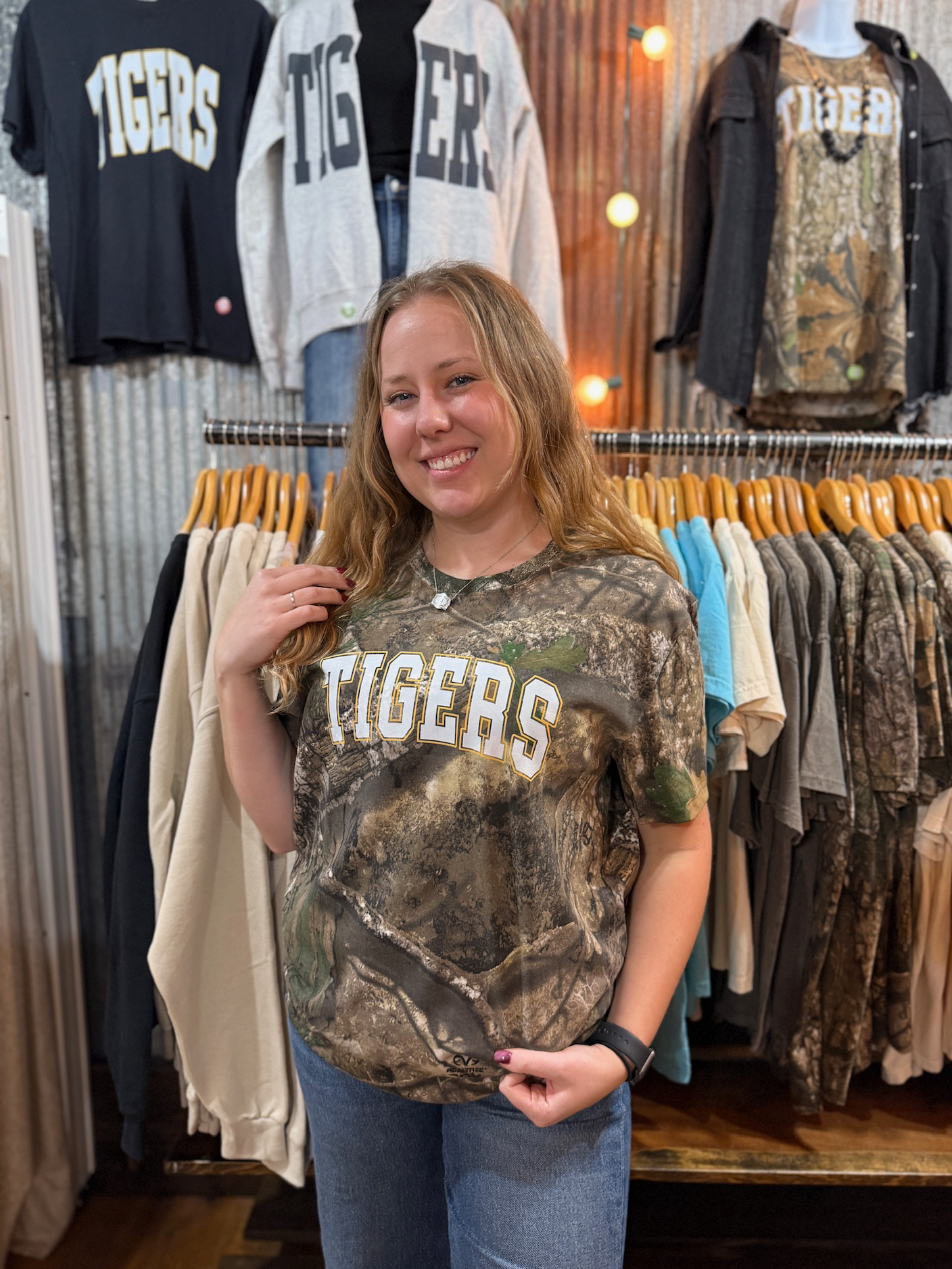 be-made-hays-ks-tigers-realtree-camo-graphic-tee Model wearing a full-length Realtree camo Tigers tee in a boutique surrounded by apparel racks.