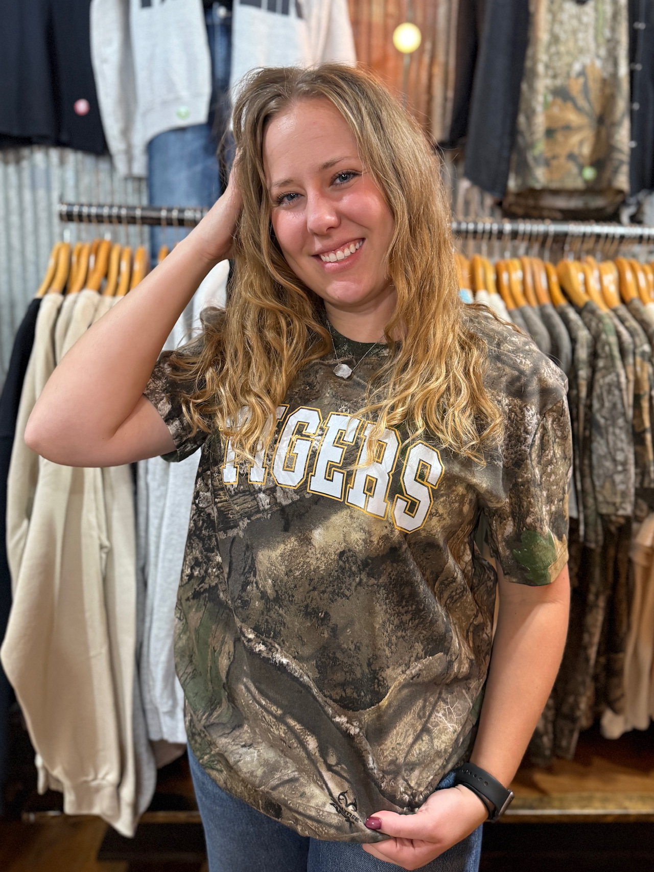 Person wearing a Realtree camo Tigers graphic tee with white and gold lettering while smiling in a boutique setting.