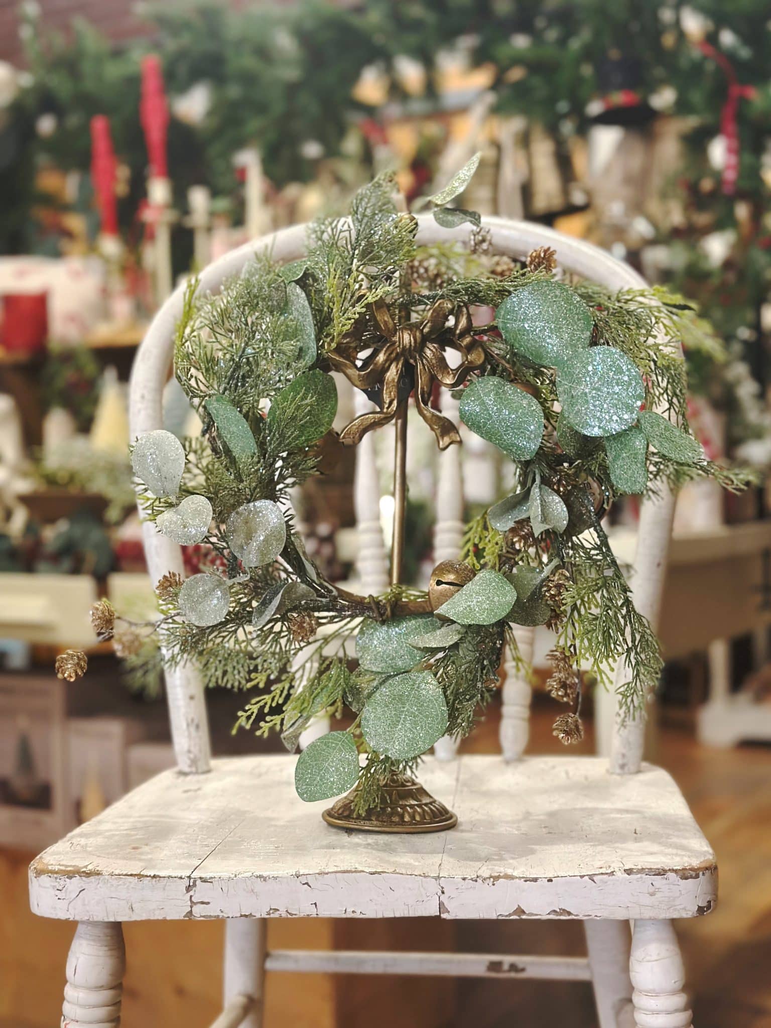 Holiday wreath with frosted cedar, eucalyptus leaves, and gold bells displayed on a gold stand atop a white vintage chair.