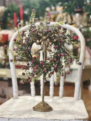 Frosted red berry wreath with green leaves displayed on an antique gold wreath stand with bow, styled on a vintage chair.