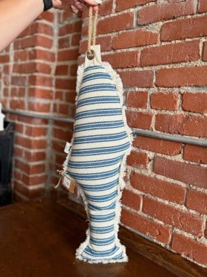 Blue Fish Shaped Pillow with blue and tan woven stripes and rope accent, styled against a brick wall.