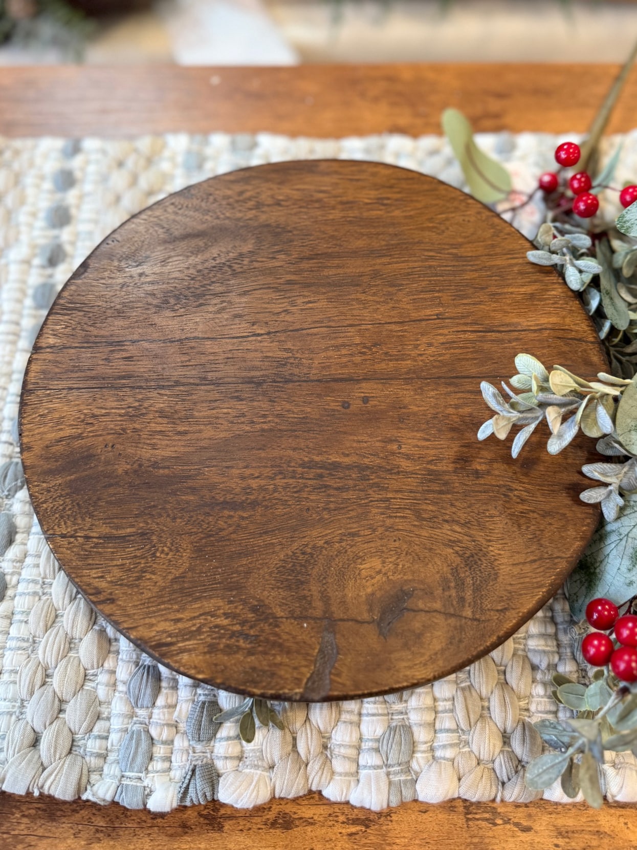 Round found wood pedestal with warm grain, styled on a woven mat with greenery and red berries.