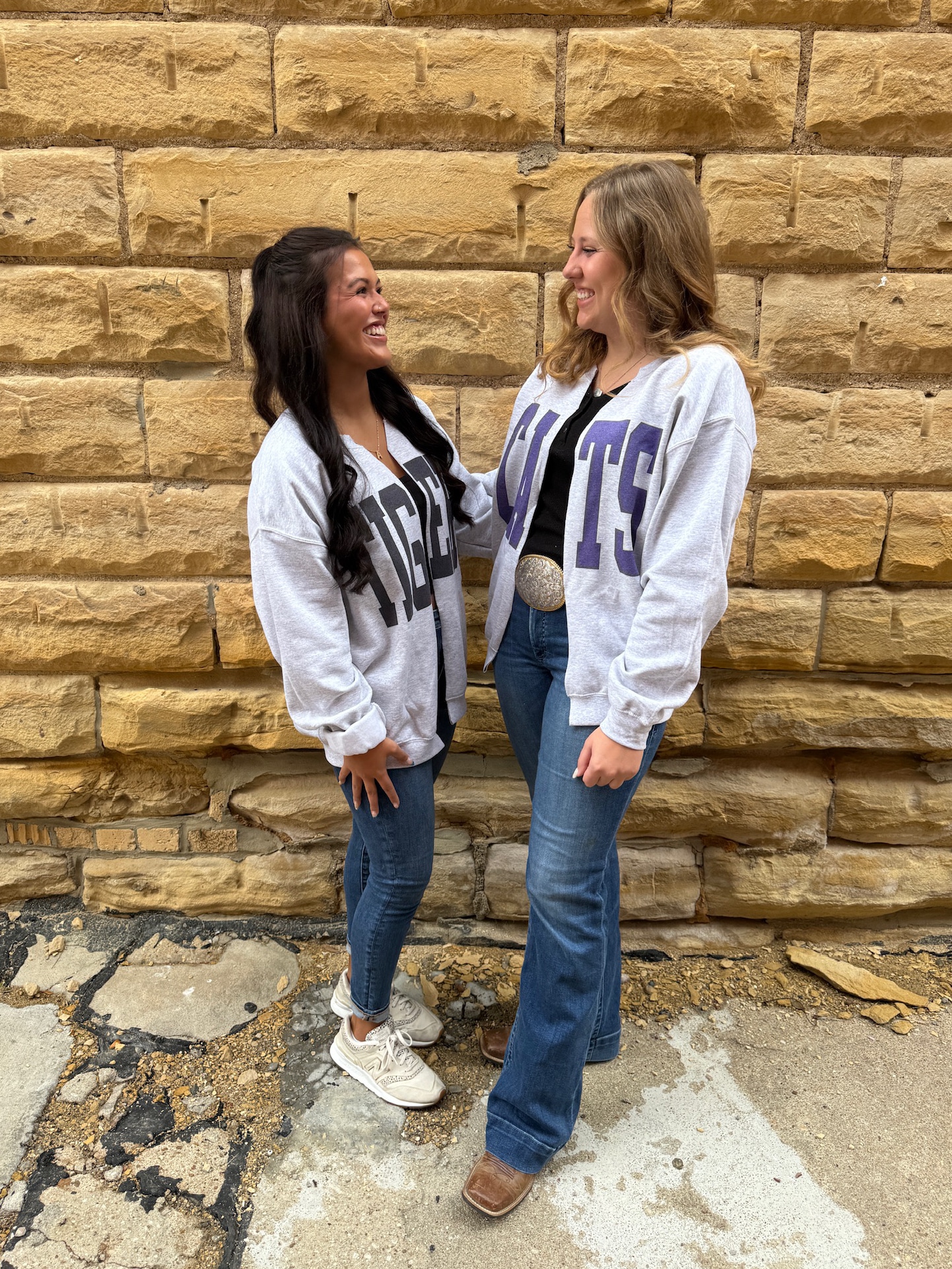 Two women wearing oversized ash gray cardigans with bold block lettering — one with “TIGERS” in black, the other with “CATS” in purple — styled casually with jeans.