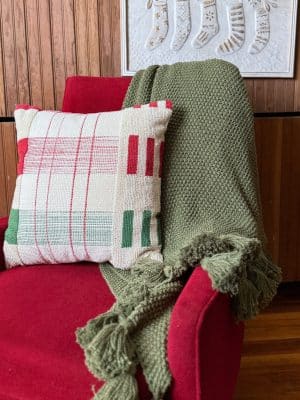 Olive green throw blanket with tassels styled on a red chair beside a patterned pillow.