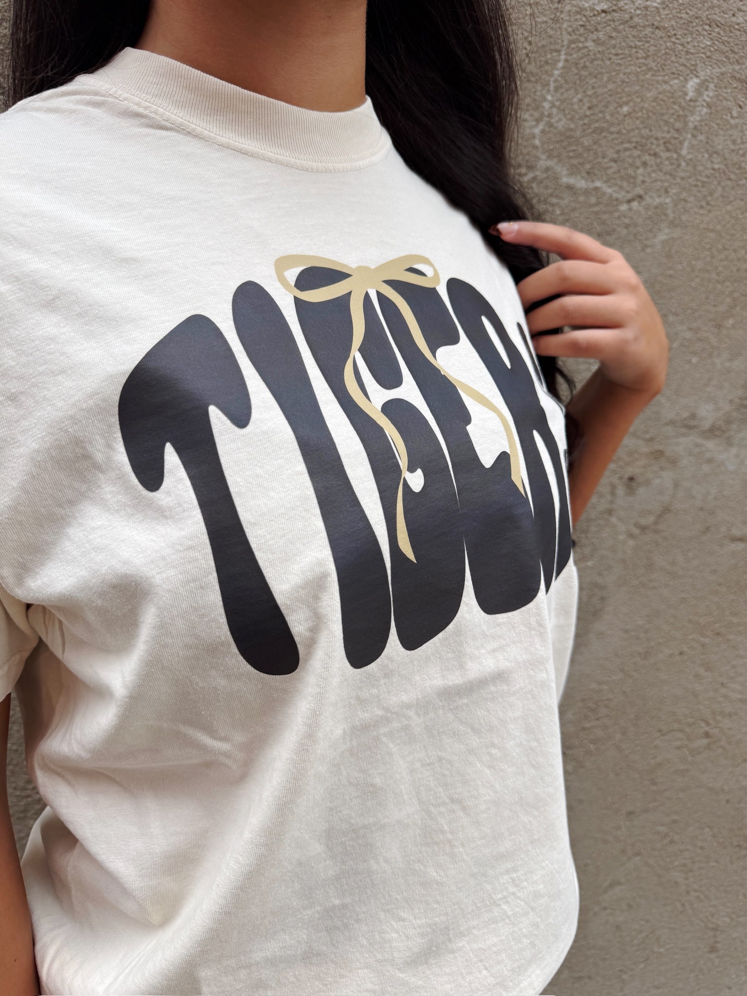 Angled shot of ivory Tigers graphic tee showing black bubble letters and gold bow print.