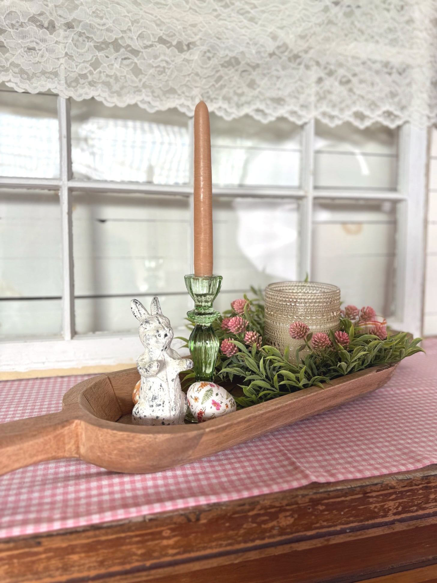 Large handcrafted wood dough bowl styled with green glass taper candle holder, distressed white bunny figurine, floral wooden eggs, greenery candle ring, champagne dotted glass candle, and pink gingham table runner.