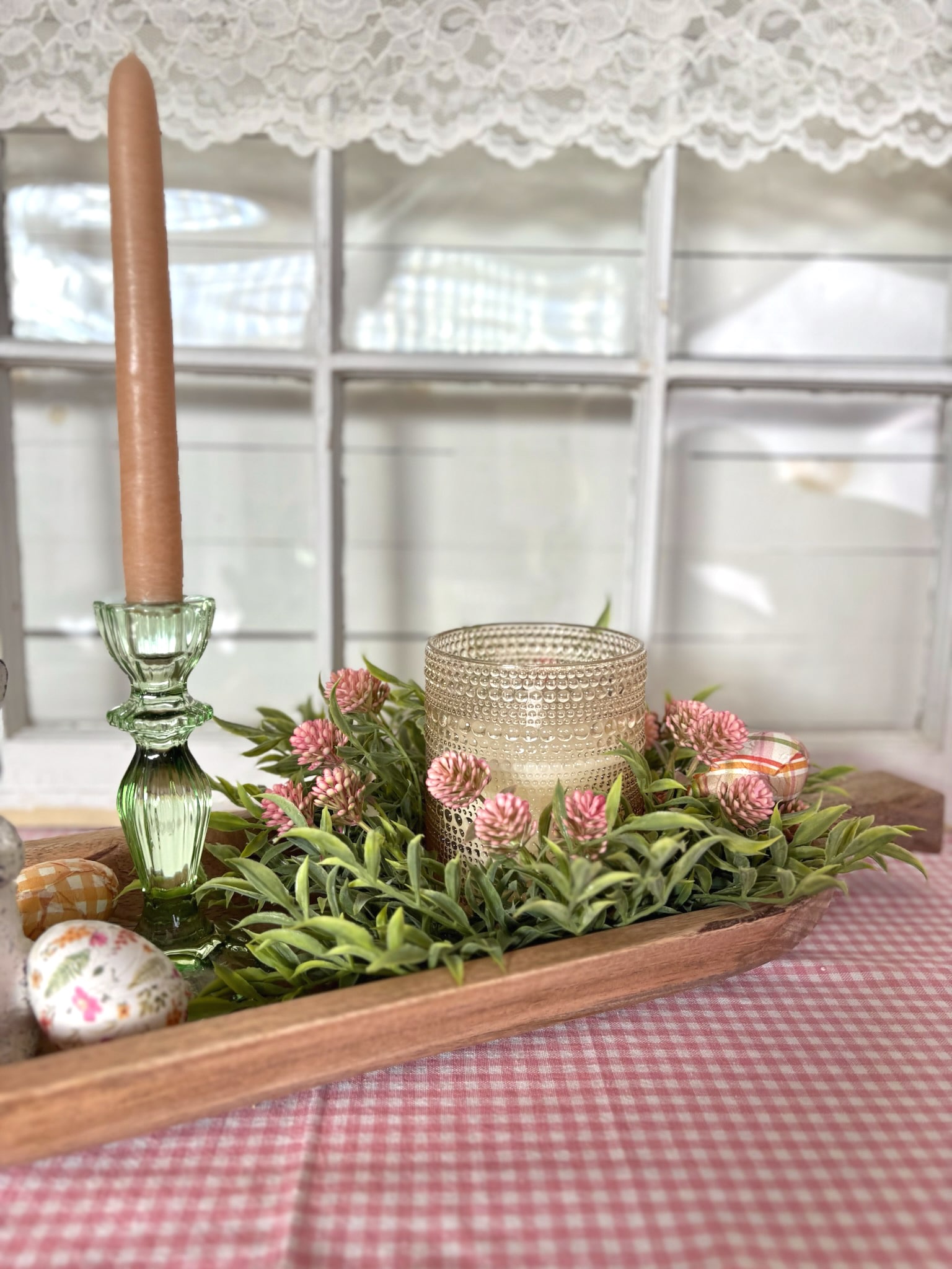 Champagne dotted glass flameless candle styled inside a large carved wood dough bowl with pink thimble buds candle ring, green glass taper candle holder, and pastel gingham wooden eggs on pink table runner.