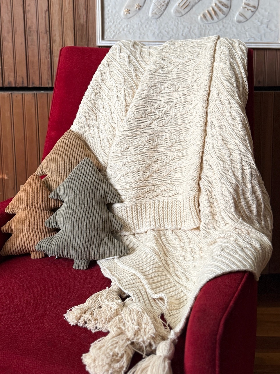 Cozy off-white cable knit throw blanket with tassels styled on a red armchair, paired with neutral corduroy tree pillows.