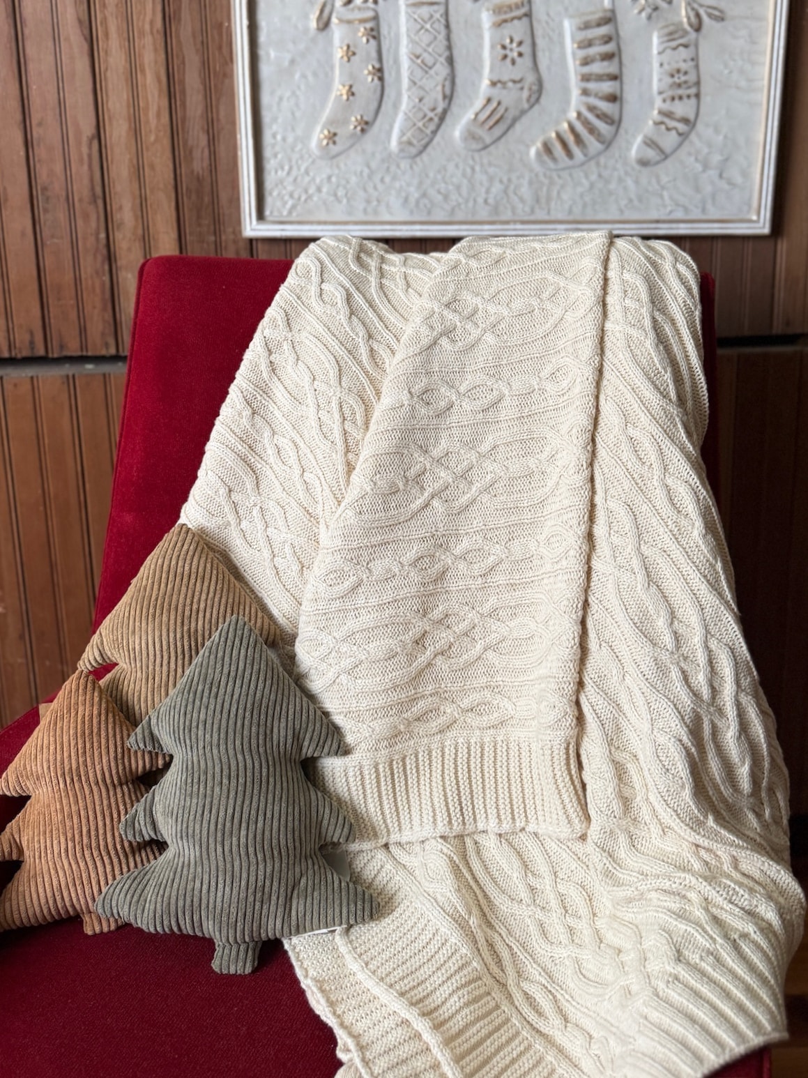Off-white cable knit throw blanket draped over a red chair with three corduroy stuffed tree pillows in neutral tones.