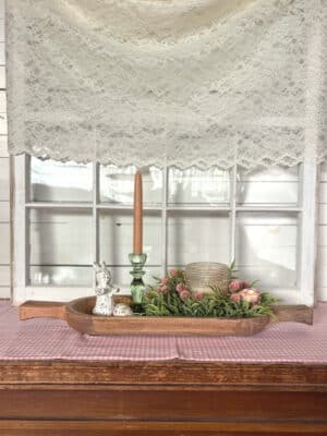 Large handcrafted wood dough bowl with handles styled with greenery, green glass taper holder, gingham eggs, white bunny figurine, pink candle ring, champagne glass flameless candle, and pink gingham table runner.