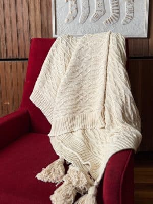 Off-white cable knit throw blanket draped over a red chair with large tassels hanging over the edge.