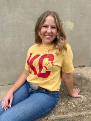 Woman wearing mustard Vintage KC Football Graphic Tee with distressed red KC football design, sitting casually outdoors.
