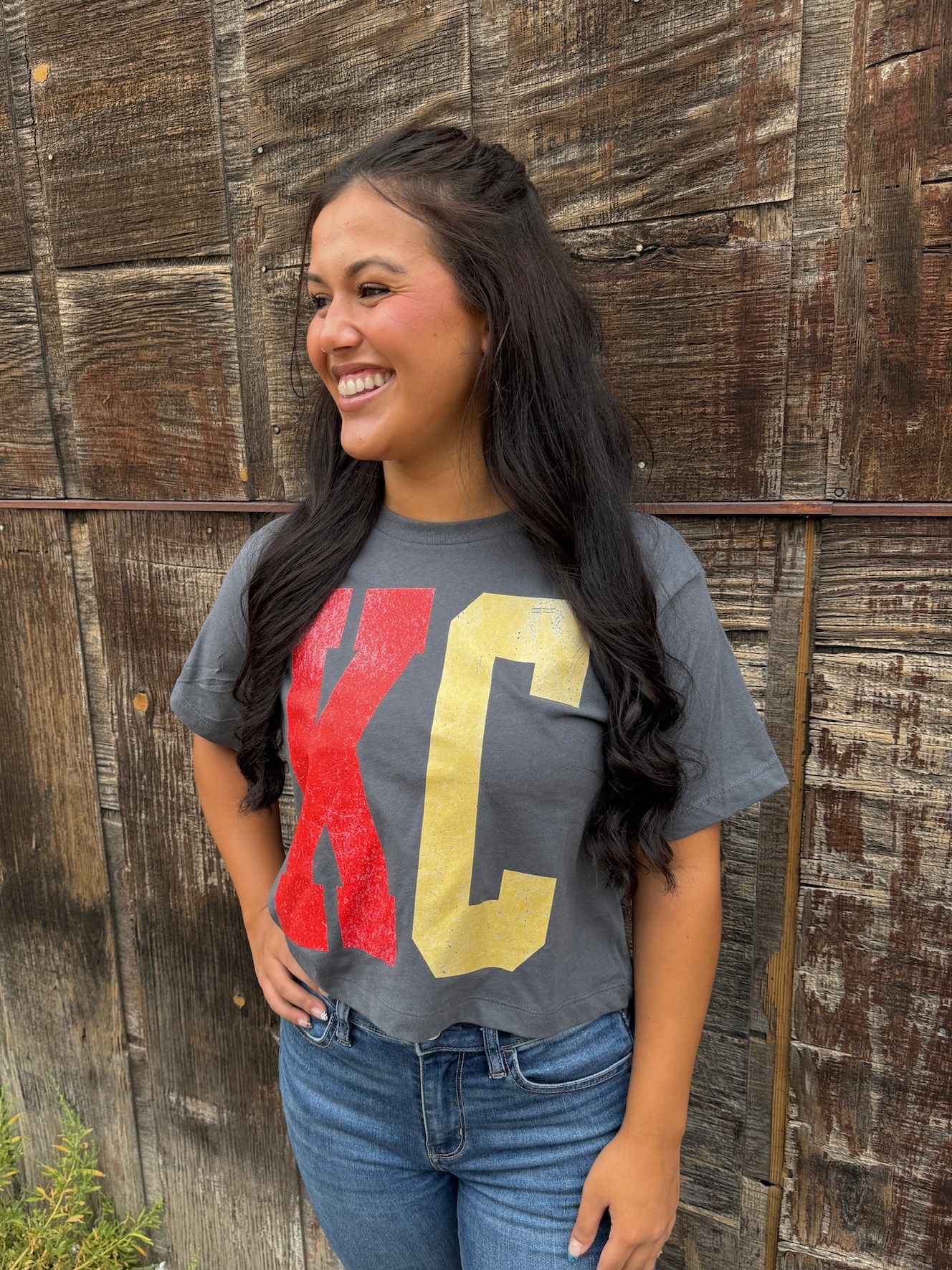 Woman smiling in cropped KC tee with distressed red and gold block lettering on pepper fabric.