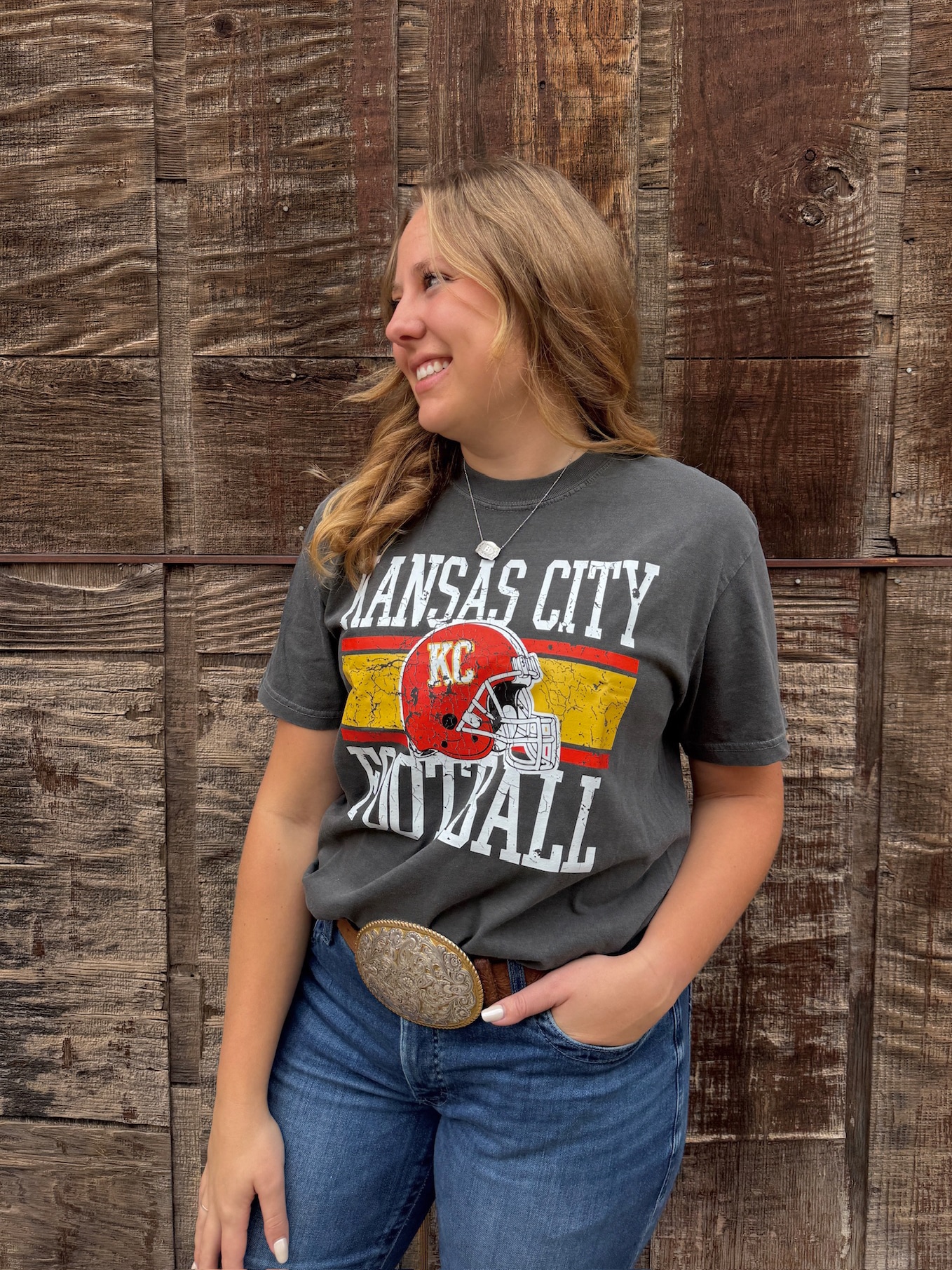 Woman smiling in pepper Kansas City Football Tee featuring distressed KC helmet and block lettering. Printed on a Comfort Colors tee in the shade Pepper, this shirt has the ultra-soft, washed cotton feel that only gets better with wear. Designed with a relaxed unisex fit, it’s perfect for pairing with denim, joggers, or leggings whether you’re tailgating at Arrowhead, watching from home, or celebrating Red Friday around town. Style: Unisex Graphic Tee Brand: Comfort Colors Color: Pepper (washed charcoal tone) Print: Distressed Kansas City Football design with red KC helmet and block lettering Fit: Relaxed, true-to-size unisex fit Material: 100% Cotton