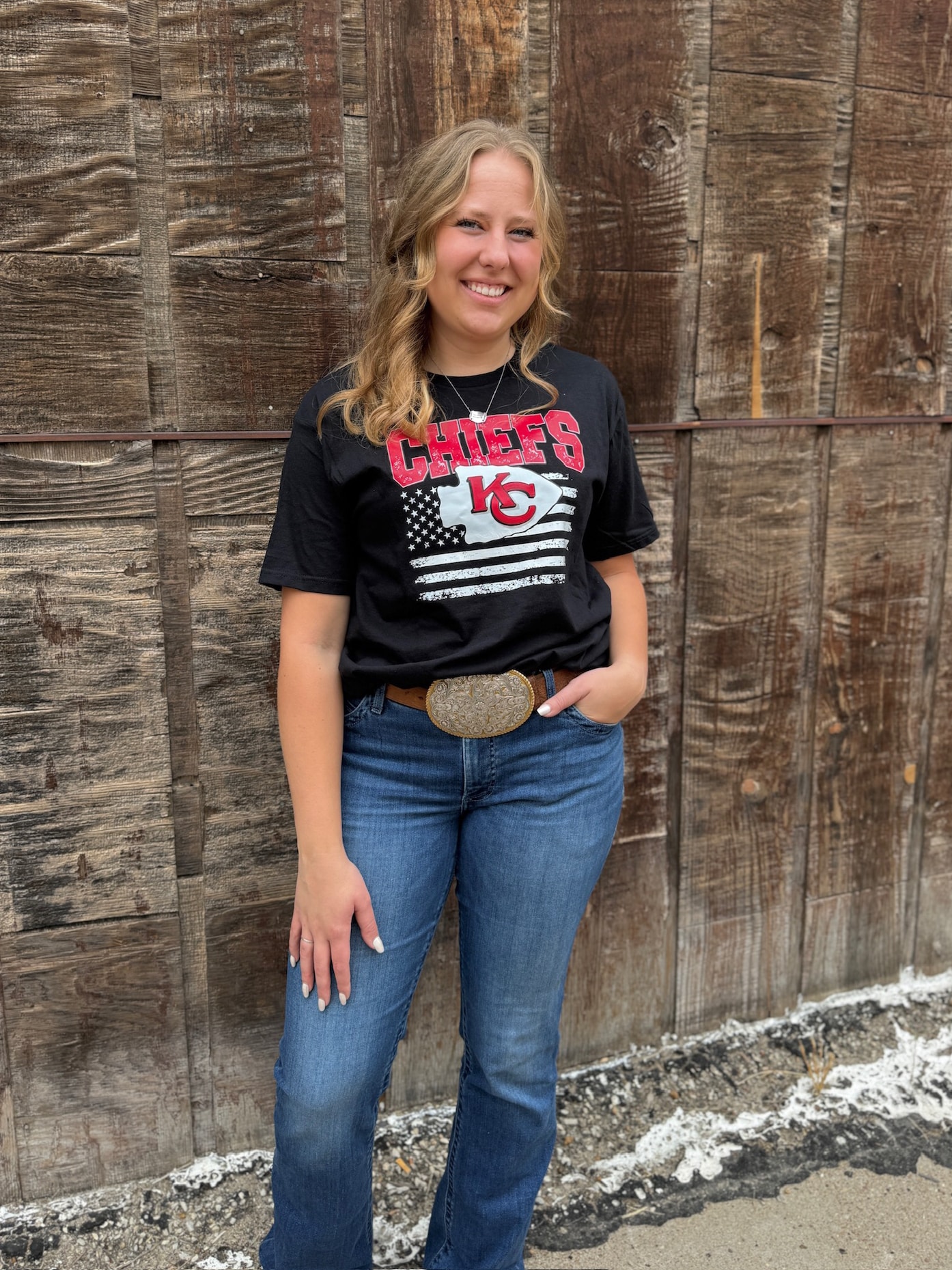 Woman wearing KC Chiefs Arrowhead Flag Graphic Tee in black with distressed red lettering and flag design.