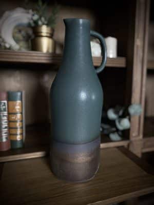 Close-up of ceramic pitcher vase with emerald green glaze and bronze base on a wooden shelf