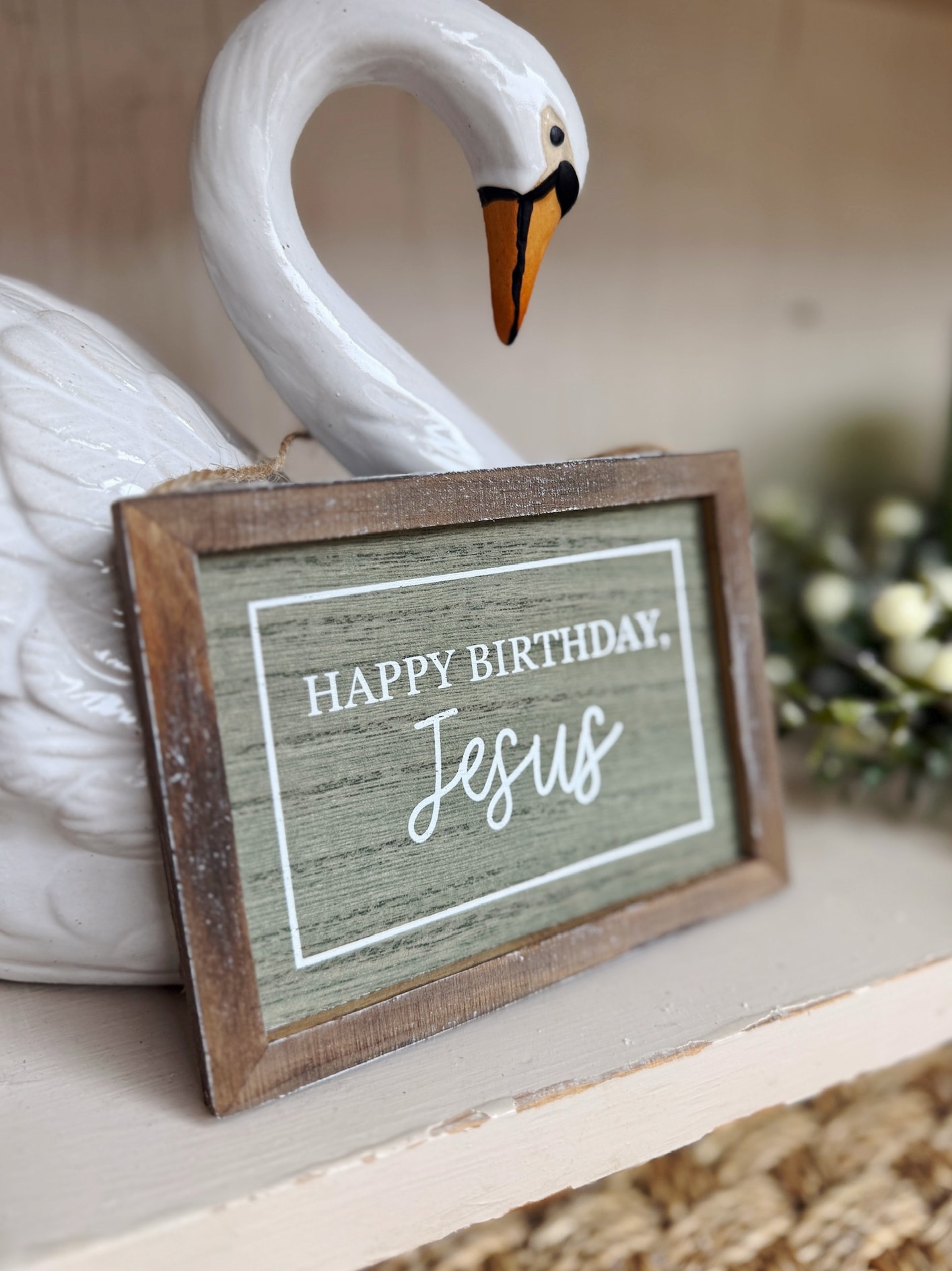 A small green and wood-framed “Happy Birthday Jesus” sign styled beside a white stoneware swan bookend.