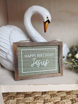 A rustic “Happy Birthday Jesus” box sign displayed in front of a white stoneware swan bookend on a shelf.