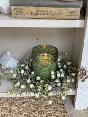 A green dotted glass flameless candle styled inside a glitter-frosted white berry leaf candle ring on a shelf.