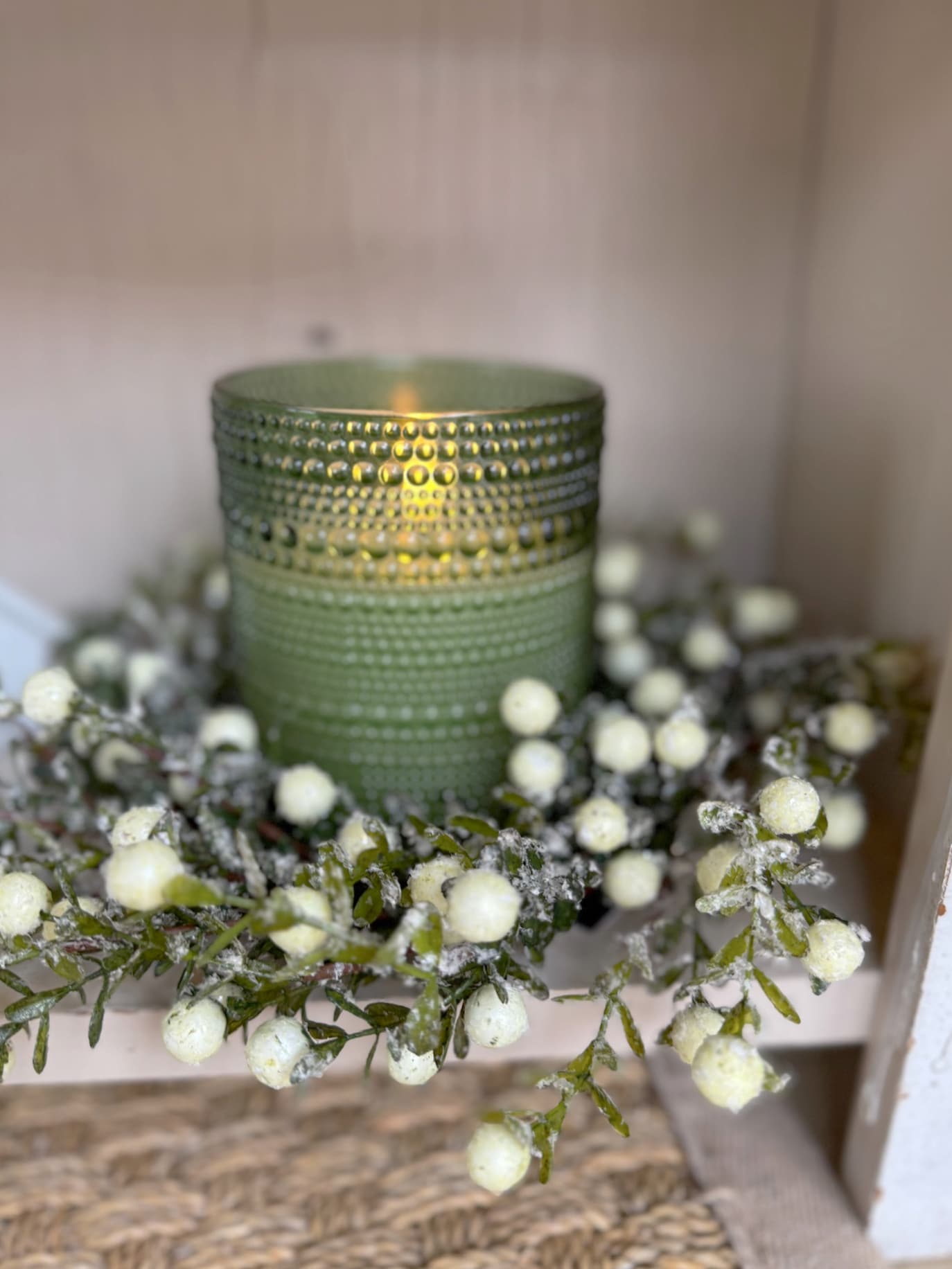 A small green dotted glass flameless candle glowing inside a frosted white berry candle ring with winter greenery.