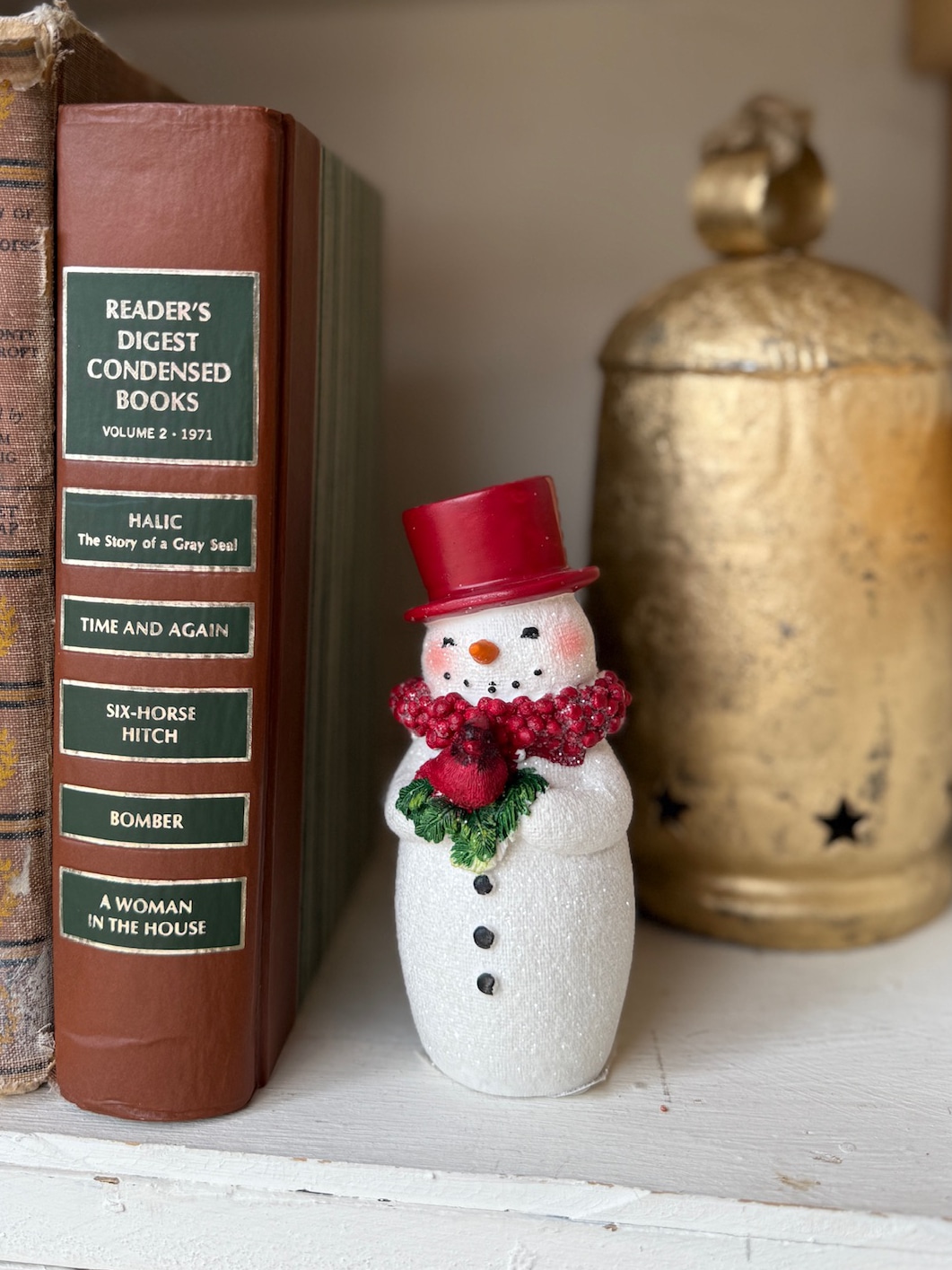 A small glittered snowman figurine wearing a red top hat and berry scarf stands beside vintage books and a gold jar, styled for Christmas décor.