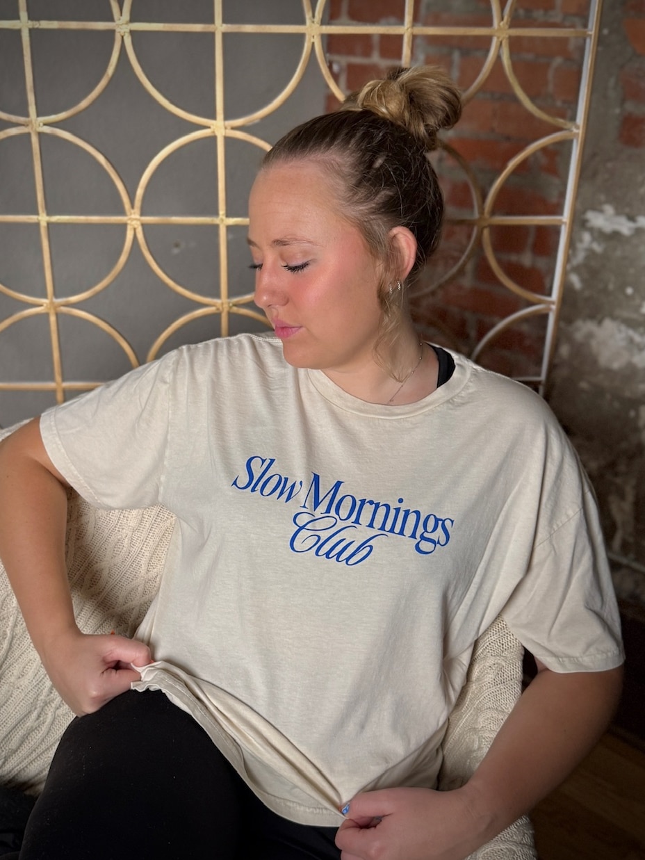 Cream unisex tee featuring “Slow Mornings Club” in blue lettering, casual fit.