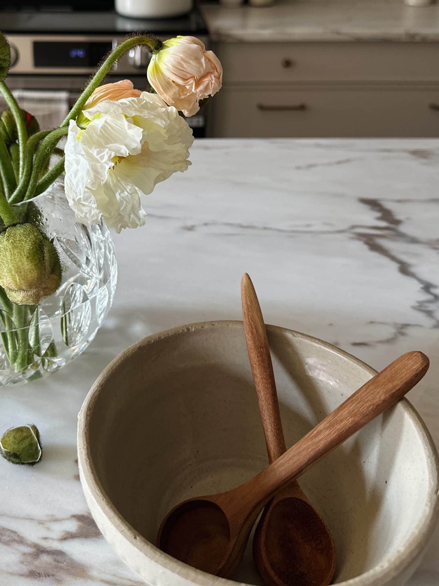 Two hand-carved wooden spoons resting in a neutral ceramic bowl on a marble countertop, next to a vase of fresh poppies
