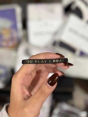 Black “You Play I Pray” silicone bracelet held in hand with blurred background.