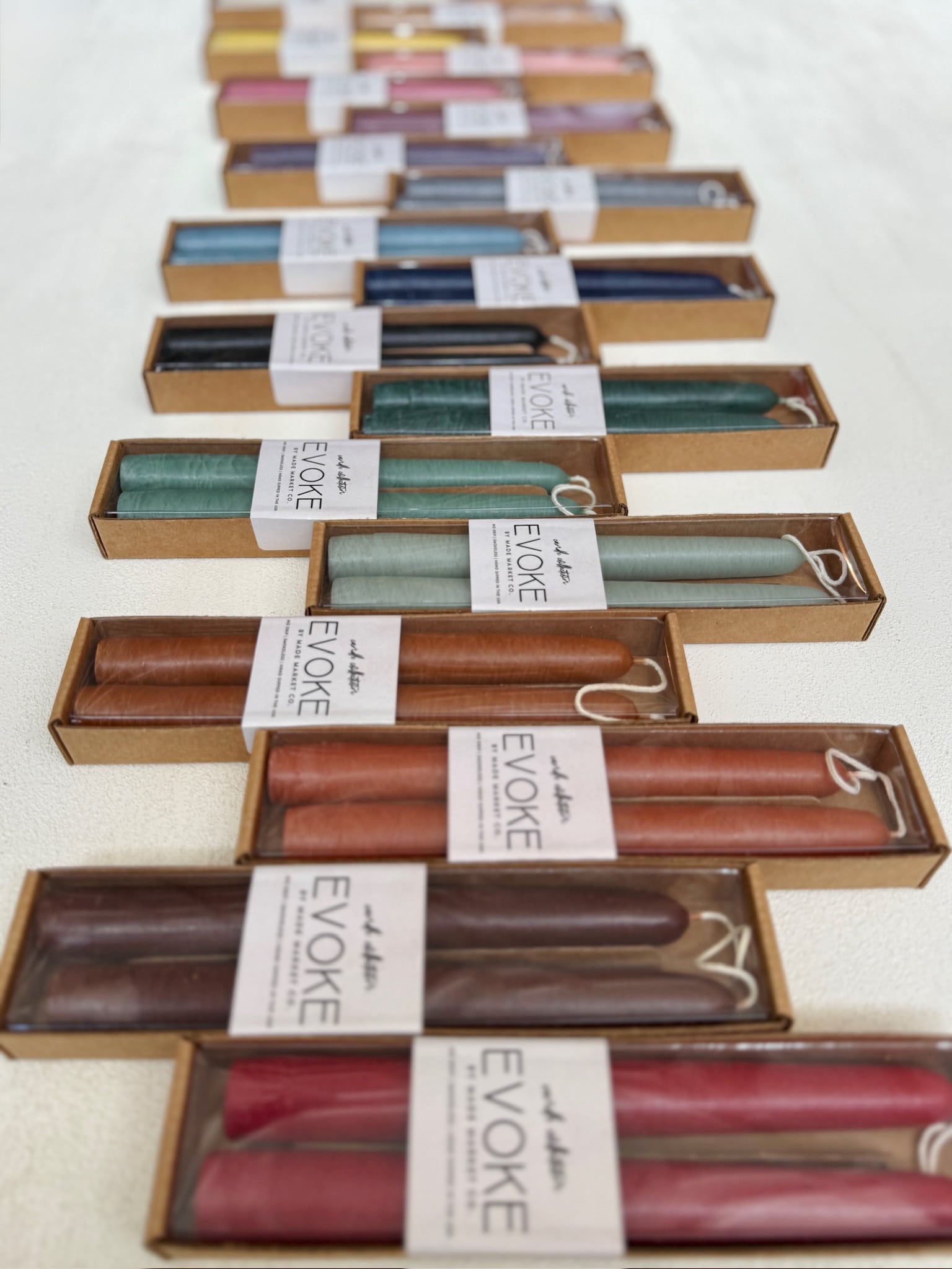 Boxes of hand-dipped Evoke taper candles in assorted colors with new white labels, arranged in rows on a white surface.