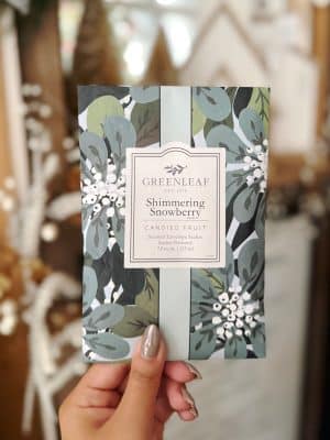 Hand holding a Shimmering Snowberry Greenleaf scented sachet in a cozy, softly blurred holiday background.