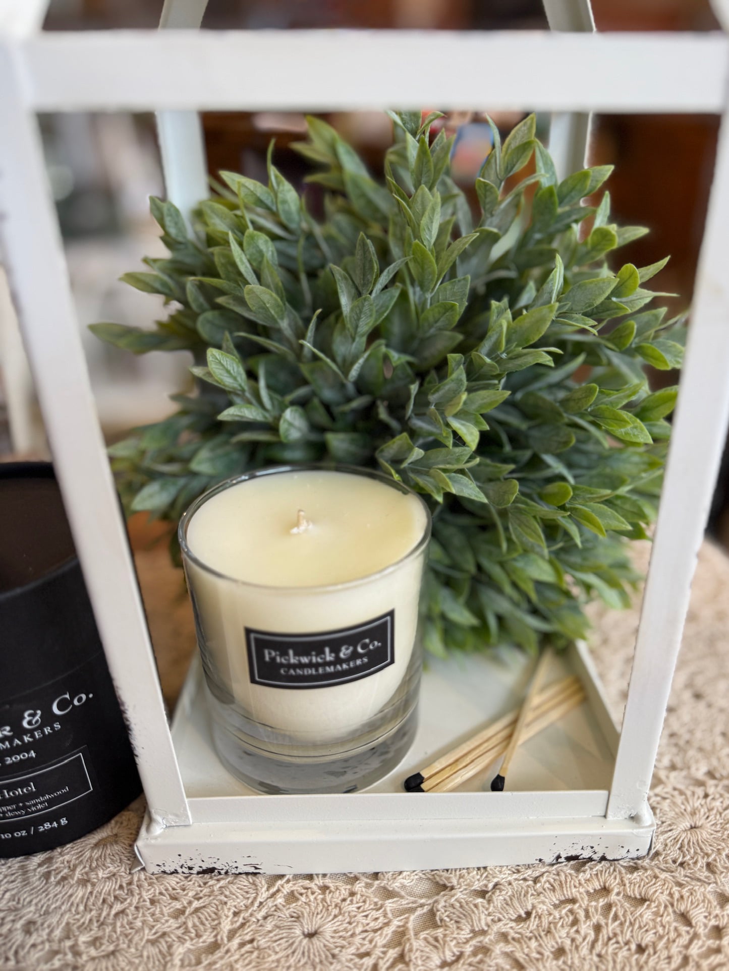 Hotel Pickwick Candle – 10 oz styled inside a distressed white metal lantern with lush green faux greenery
