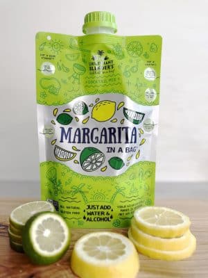 Margarita Cocktail in a Bag mix displayed upright on a wood surface, surrounded by sliced lemons and limes.