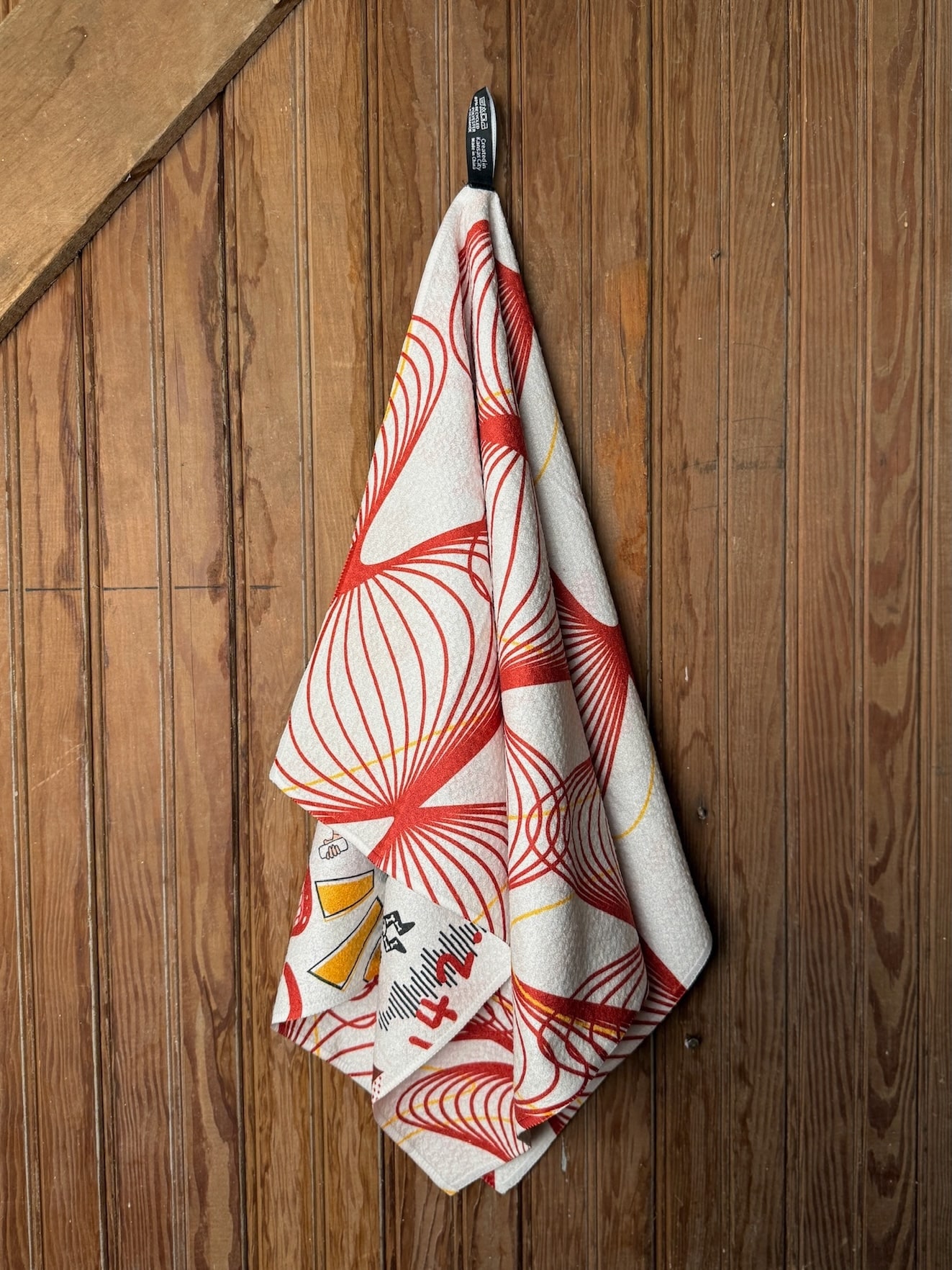 Microfiber KC Chiefs tea towel with colorful red, yellow, and white illustrations including football icons, hearts, and game-day phrases, shown flat and hanging on wood wall.