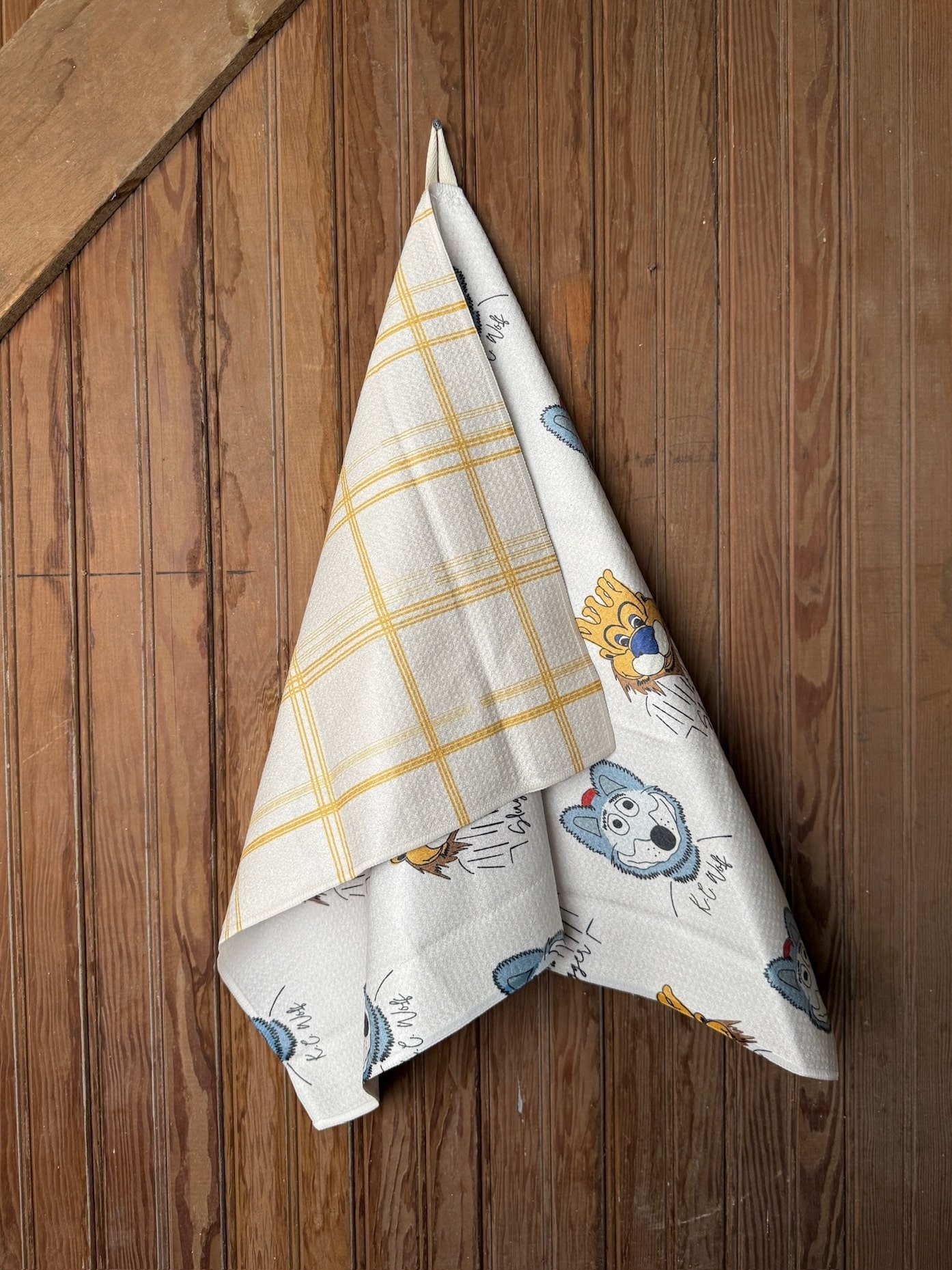 Microfiber tea towel with KC Wolf and Royals Sluggerrr mascot illustrations, shown folded and hanging on rustic wooden wall.