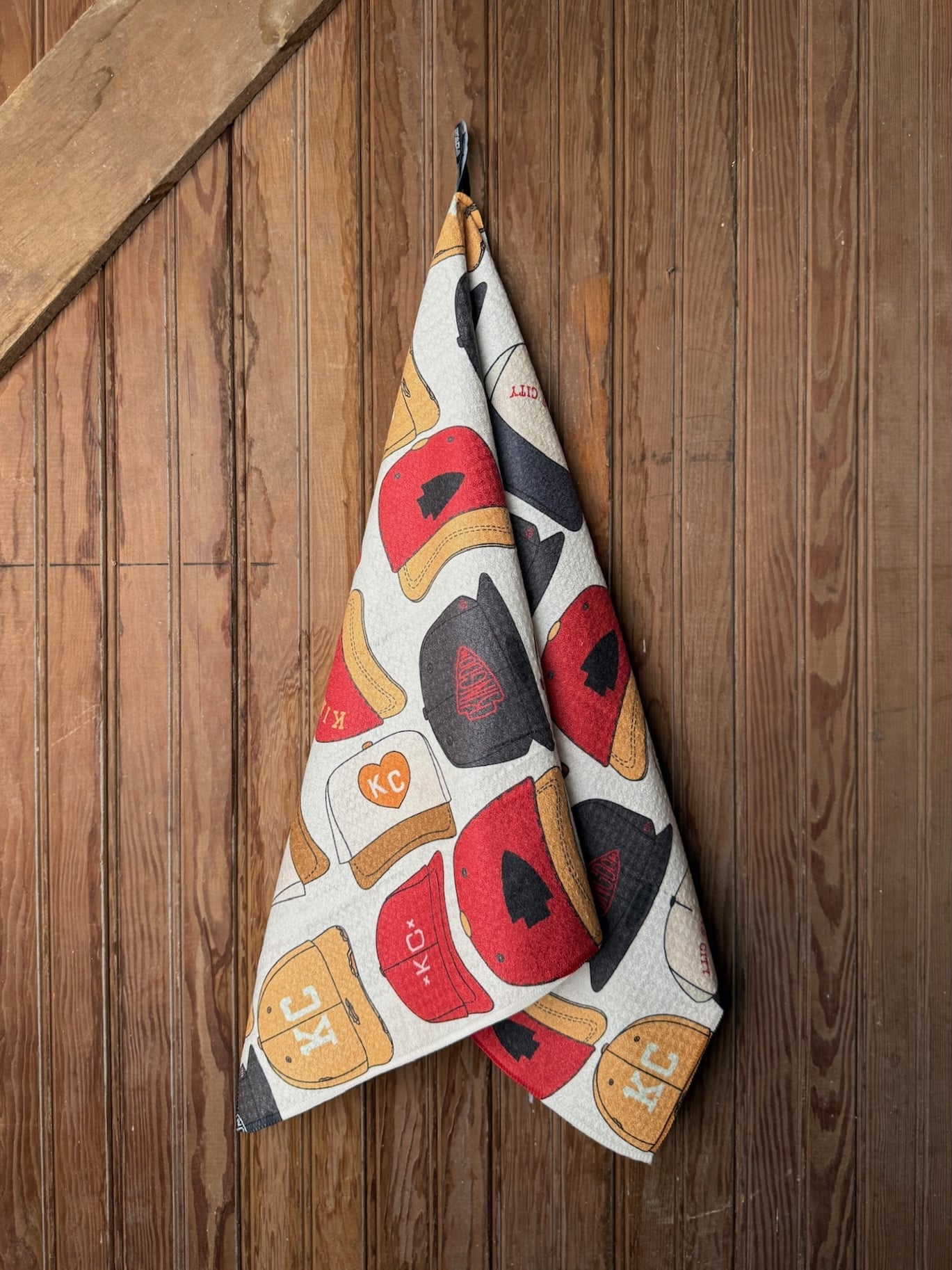be-made-hays-ks-kingdom-hats-kc-chiefs-tea-towel Double-sided Chiefs Kingdom Hats Tea Towel with a mix of KC and Kingdom hat designs in red, gold, and black, photographed hanging on a wood wall.