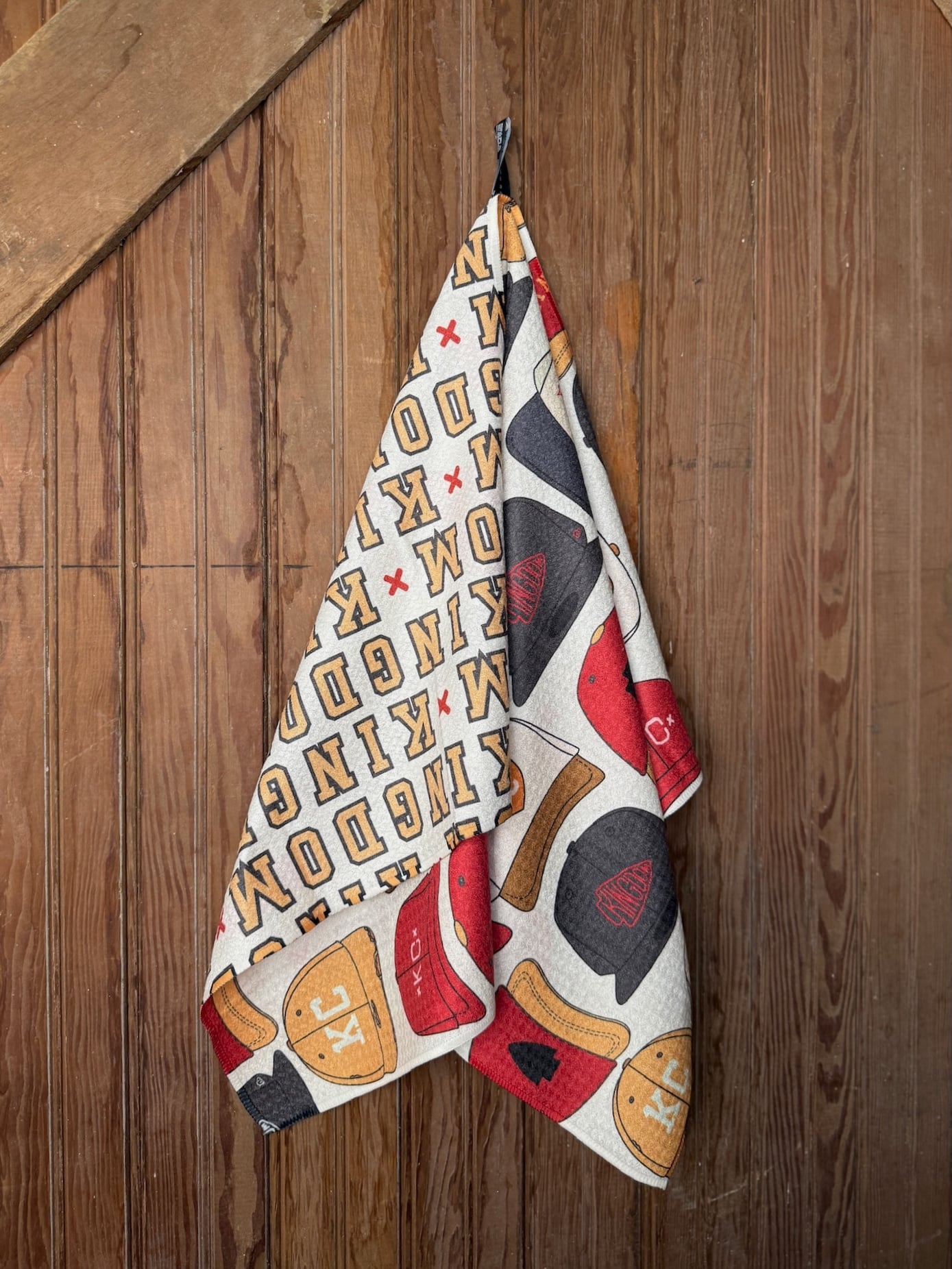 Microfiber KC Chiefs Hats Tea Towel with red, gold, and black hat pattern, shown flat and hanging against a rustic wooden surface.