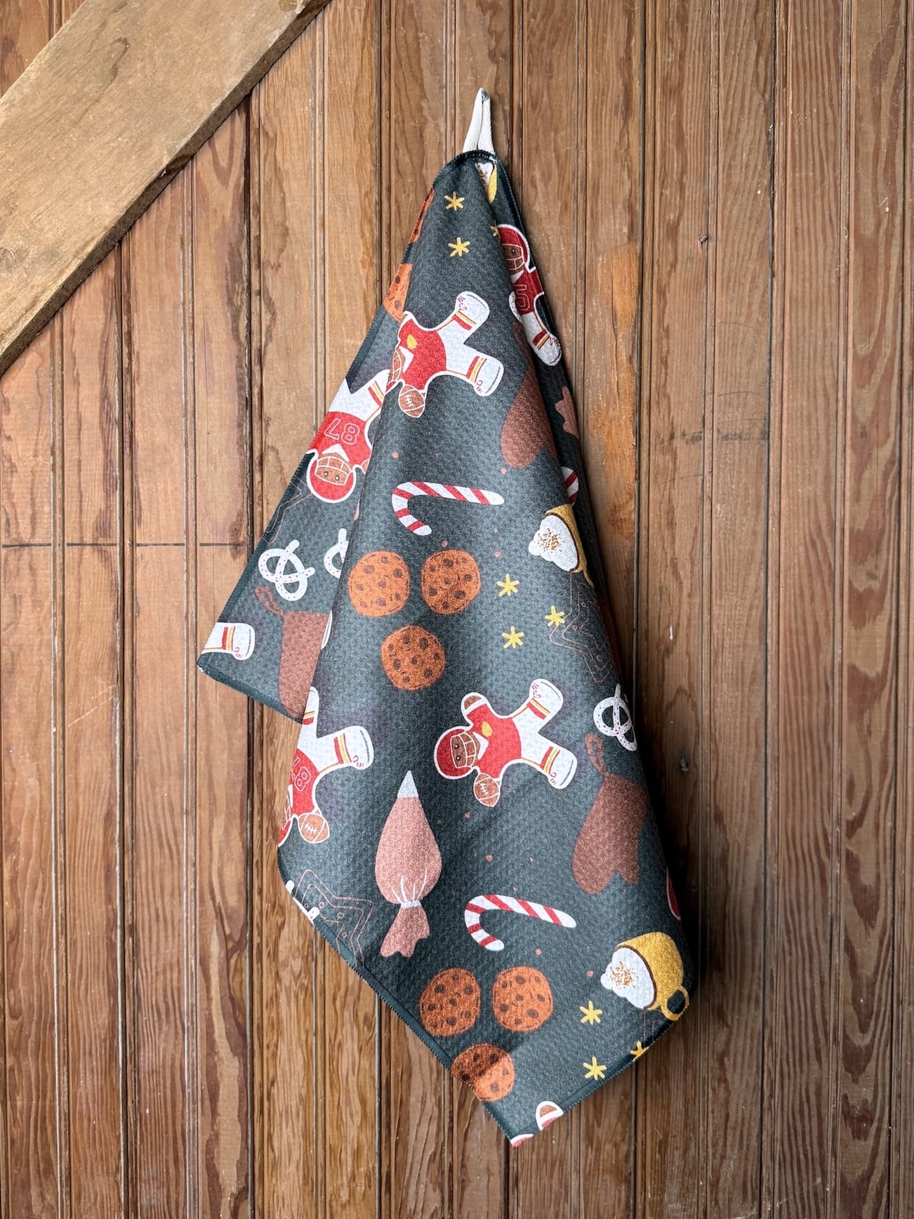 made-market-co-hays-ks-chiefs-christmas-cookies-kitchen-towel Holiday tea towel decorated with red and gold gingerbread players, cookies, and candy canes on dark waffle fabric, folded on wood surface.