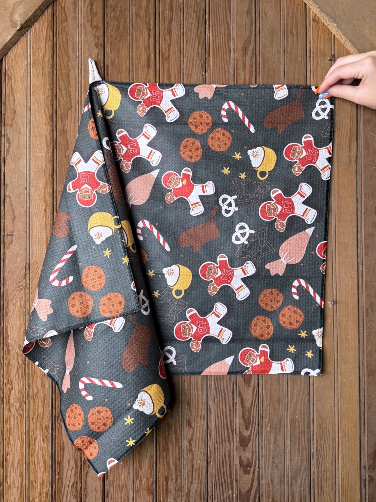Chiefs Christmas Cookies Tea Towel featuring gingerbread football players, candy canes, and cookies on a dark gray background, hanging on a wood wall.