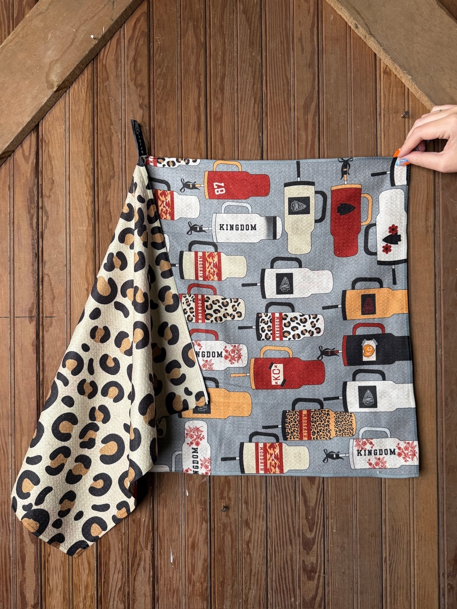 Double-sided KC Chiefs tea towel with tumbler illustrations and bold leopard print design, displayed hanging on rustic wooden background.