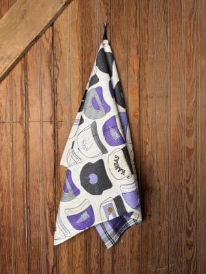 Double-sided K-State Wildcats Hats Tea Towel featuring purple and gray hat illustrations with Wildcat lettering, shown hanging neatly on a wood panel wall.
