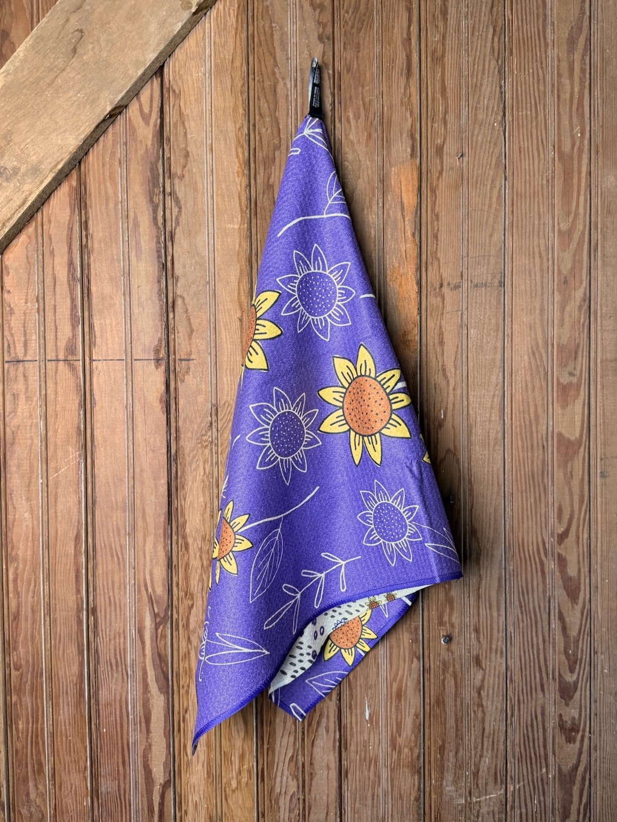 KSU Tailgate Tea Towel featuring tailgate illustrations, fans, and “Eat ’Em Up” lettering with a sunflower pattern on the reverse, displayed hanging on wooden wall.