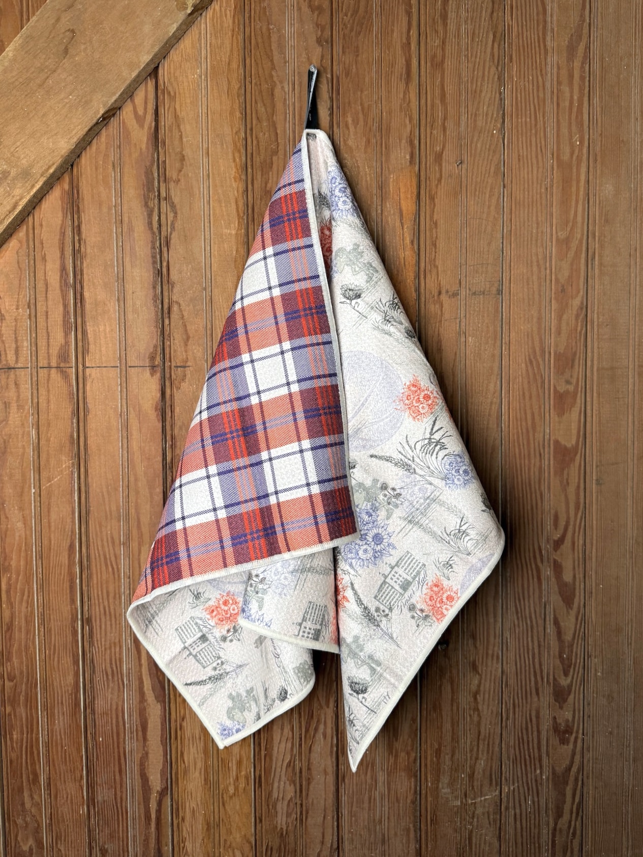 Double-sided KU 1912 Basketball Tea Towel hanging at an angle showing the vintage print front and plaid reverse on a rustic wood backdrop.