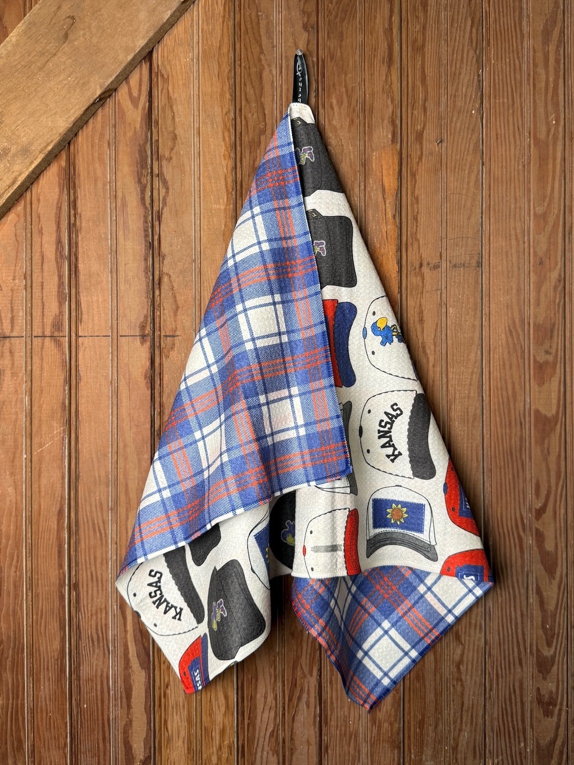 KU Hats Tea Towel draped on a wooden backdrop showing the vintage hat print front and plaid reverse with Jayhawk colors.
