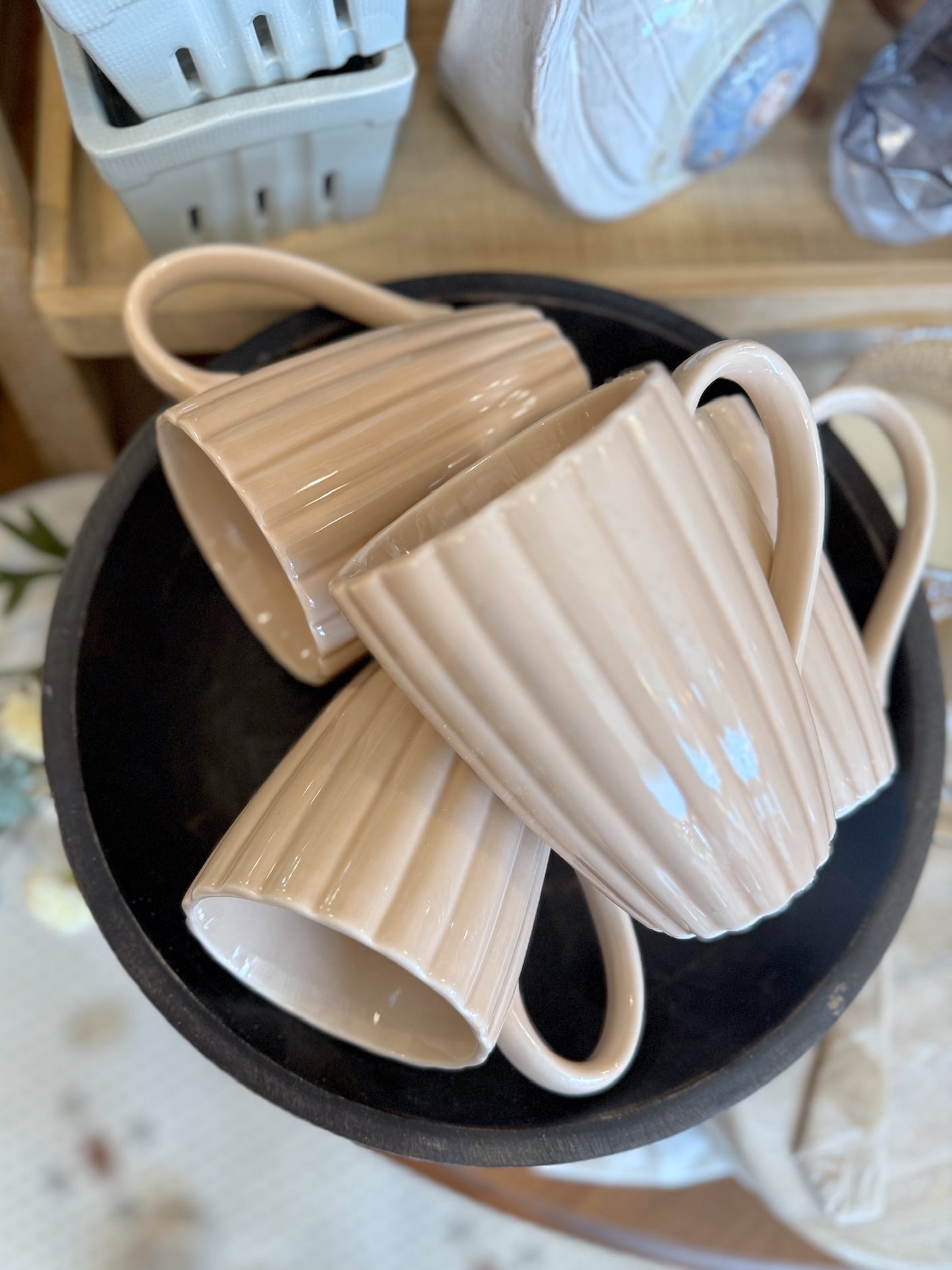 Three cream ribbed stoneware mugs stacked together in a black bowl on a wooden shelf display.