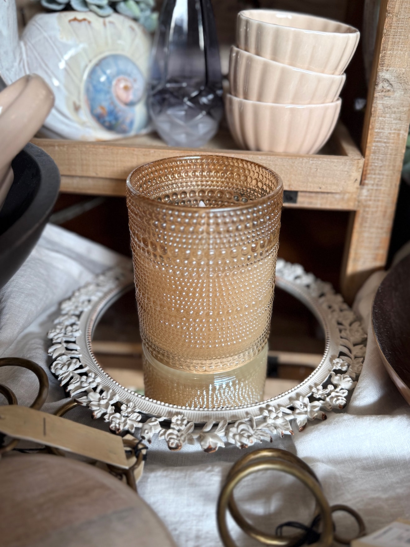 Textured champagne dotted flameless candle displayed on white floral framed mirror tray surrounded by home accents.