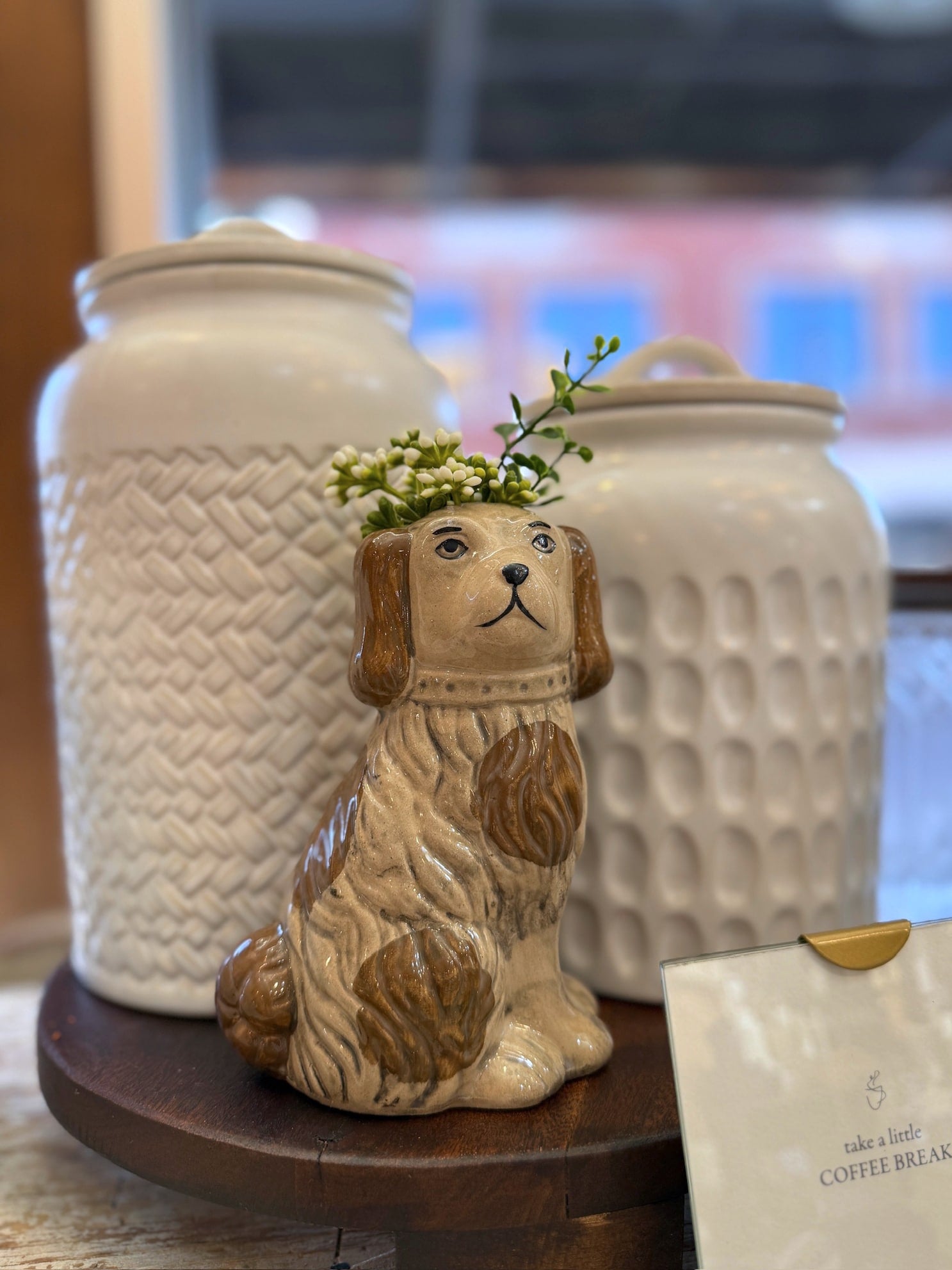 Close-up of Staffordshire-style dog planter with greenery inside, displayed beside white pottery jars near a window.
