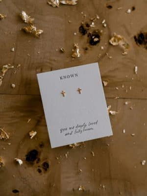 Minimal 14k gold-filled cross stud earrings on a cotton letterpress card reading “you are deeply loved and fully known,” styled with warm gold flecks.