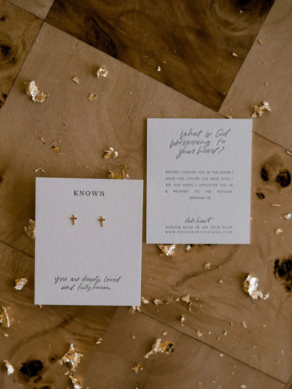 Dainty 14k gold-filled cross stud earrings displayed on a cotton letterpress card with faith-based wording, styled flat with scattered gold leaf accents.