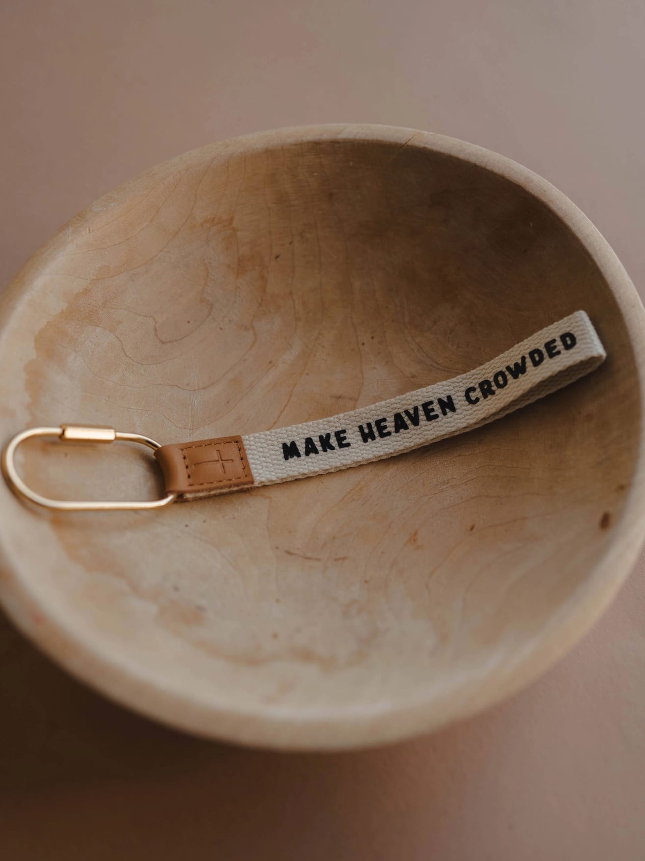 Neutral keychain wristlet with “Make Heaven Crowded” text, leather cross tag, and gold carabiner clip.