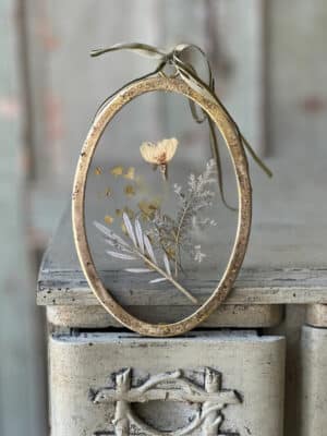 Distressed gold oval frame with pressed greenery and florals displayed on a shelf with velvet ribbon hanger.